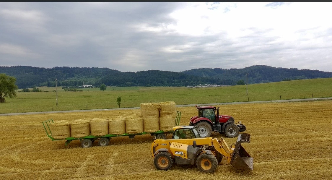 Traktor s prívesom naloženým balíkmi slamy a žltý teleskopický nakladač na strnisku
